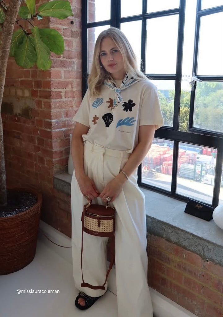Woman wearing The Artist Series TBCo x Chiara Perano Cotton T-Shirt with abstract shapes, white trousers, and a patterned scarf, holding a woven handbag indoors.