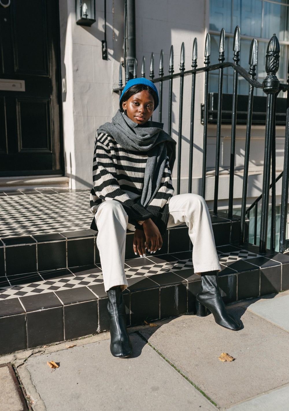 A person wearing TBCo Lambswool Blanket Scarf in Charcoal Melange styled with striped jumper, white trousers and blue beanie on steps