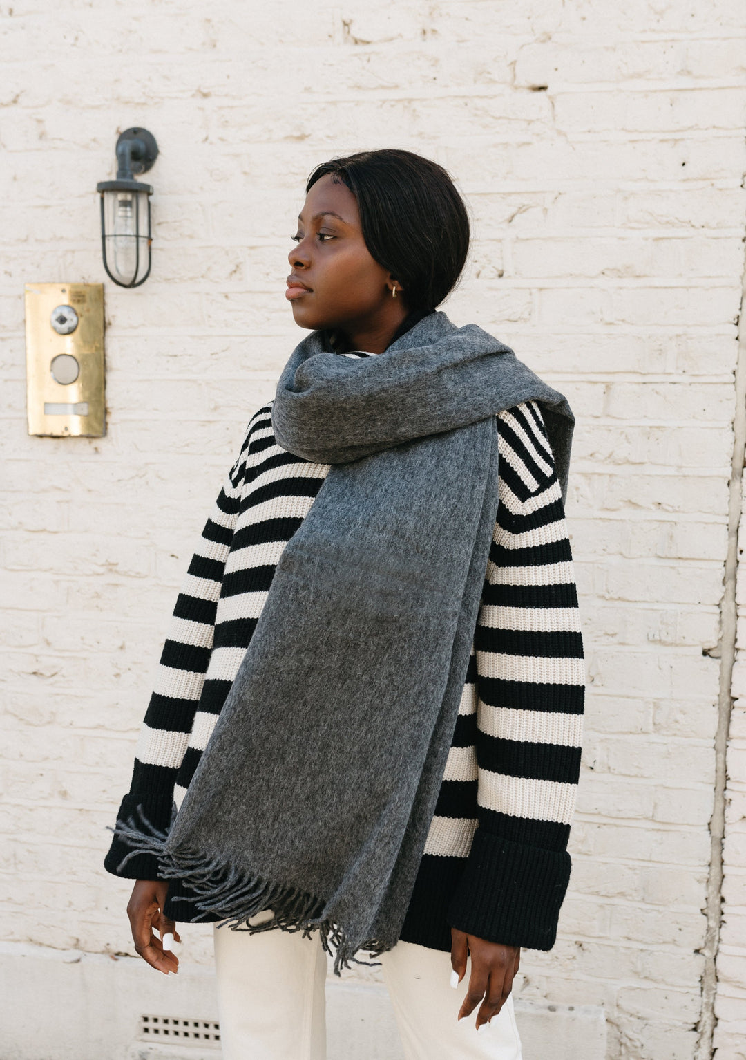TBCo Lambswool Blanket Scarf in Charcoal Melange styled with striped jumper against white brick wall, showing luxurious drape and fringe