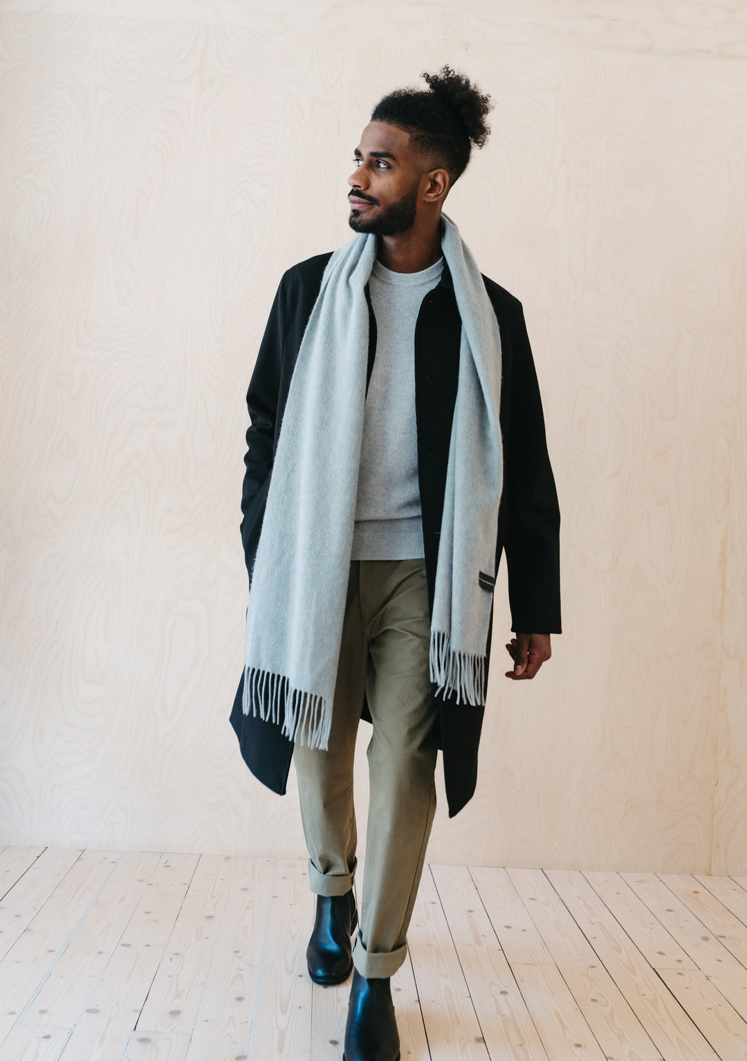 A person wearing TBCo Men's Cashmere Scarf in Light Grey Melange styled with black coat and grey trousers against neutral backdrop