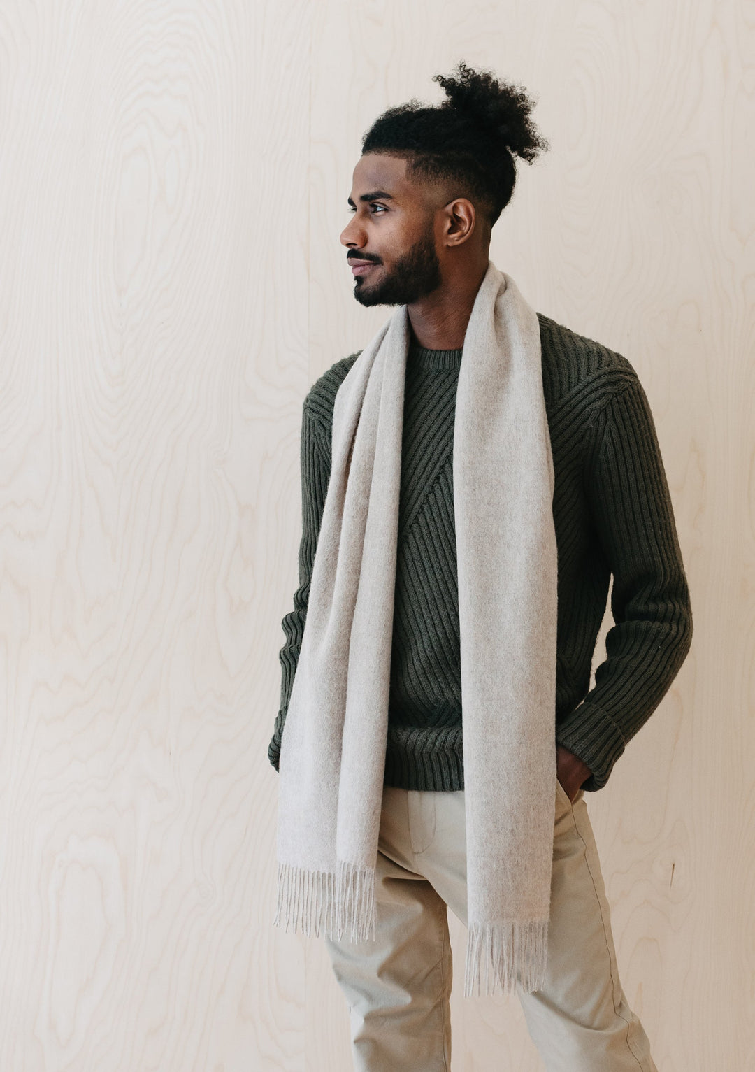 A person wearing TBCo Men's Lambswool Scarf in Oatmeal Melange draped over a dark green knit jumper, showcasing the soft fringed texture