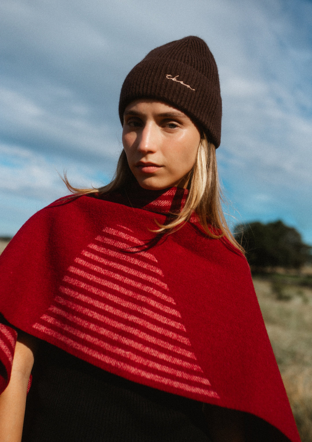 Woman wearing a TBCo Cashmere & Merino Beanie in Chocolate outdoors with a red cape against a blue sky background