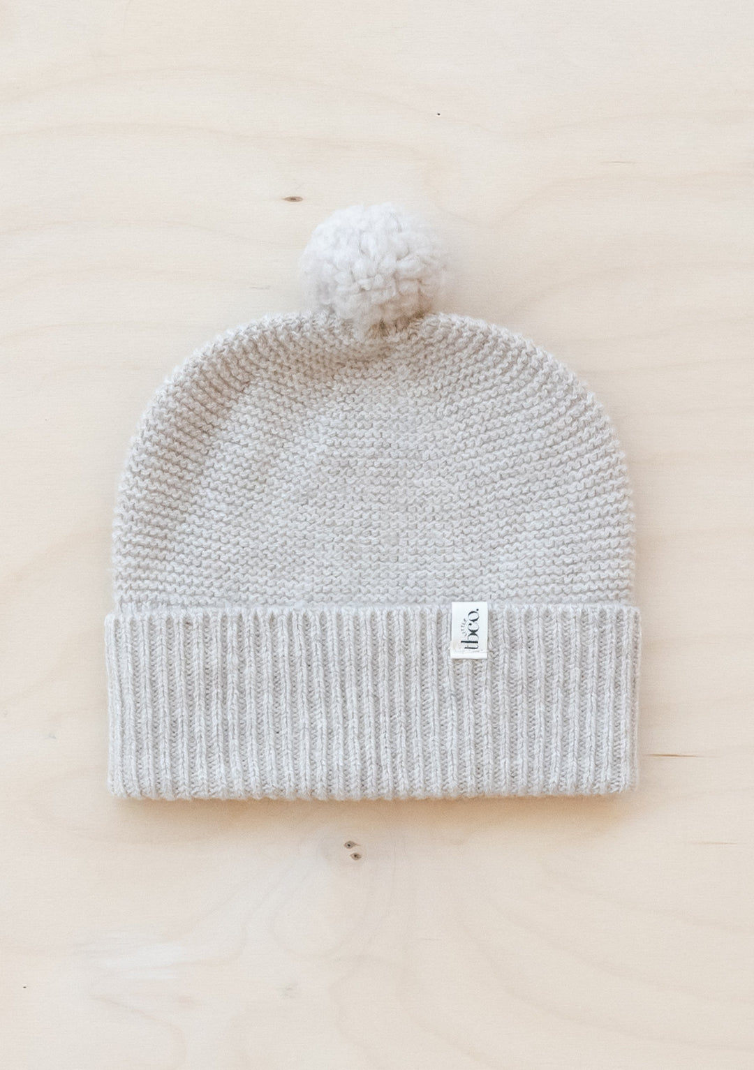 TBCo Merino Wool Baby Beanie in oatmeal melange with white pom-pom, knitted ribbed hem and small brand label