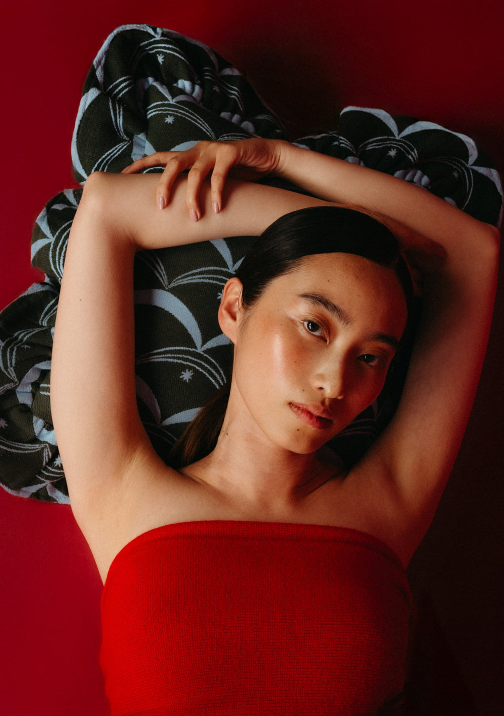 Young woman with dark hair reclines on a patterned TBCo Cotton Cushion Cover in Green Circus, featuring a bold print and deep frill, set against a red cirus inspired background.