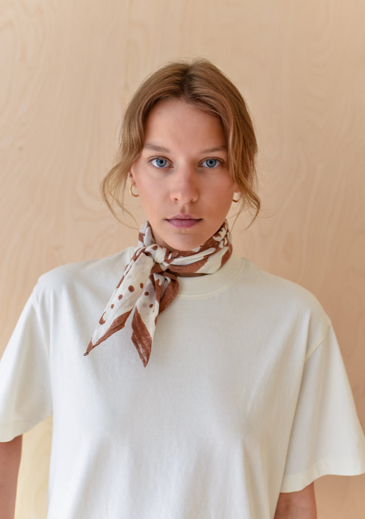 Young woman wearing The Artist Series TBCo x Frankie Penwill Cotton Scarf, cream with brown accents, tied around neck, against light wood-paneled background.