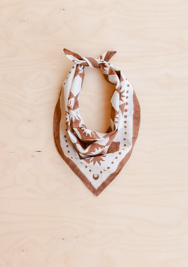 The Artist Series TBCo x Frankie Penwill Cotton Scarf, featuring stylized suns and moons, knotted into a kerchief on a wooden surface.