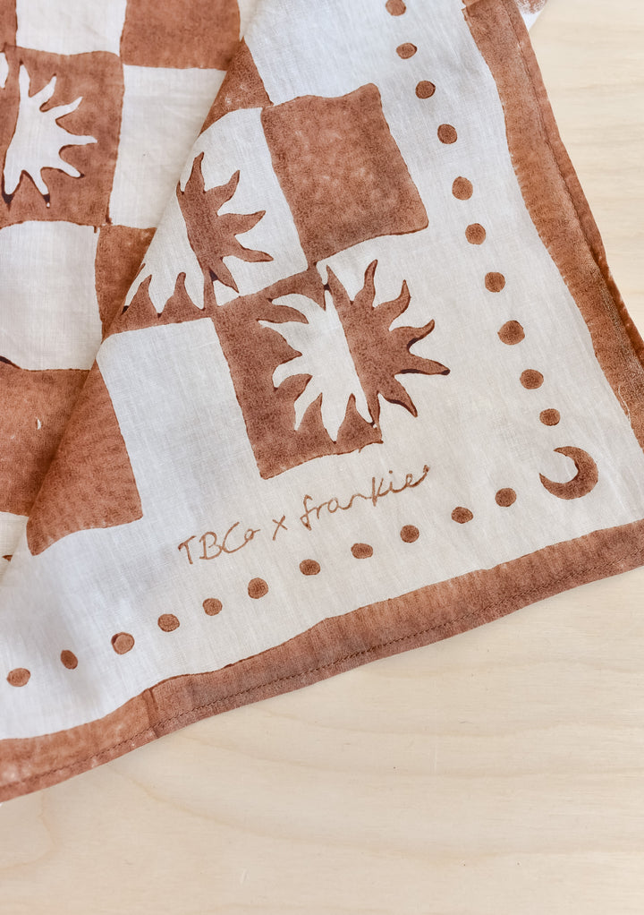 The Artist Series TBCo x Frankie Penwill Cotton Scarf features a rust-brown sunburst design on off-white, with a crescent moon in the corner.