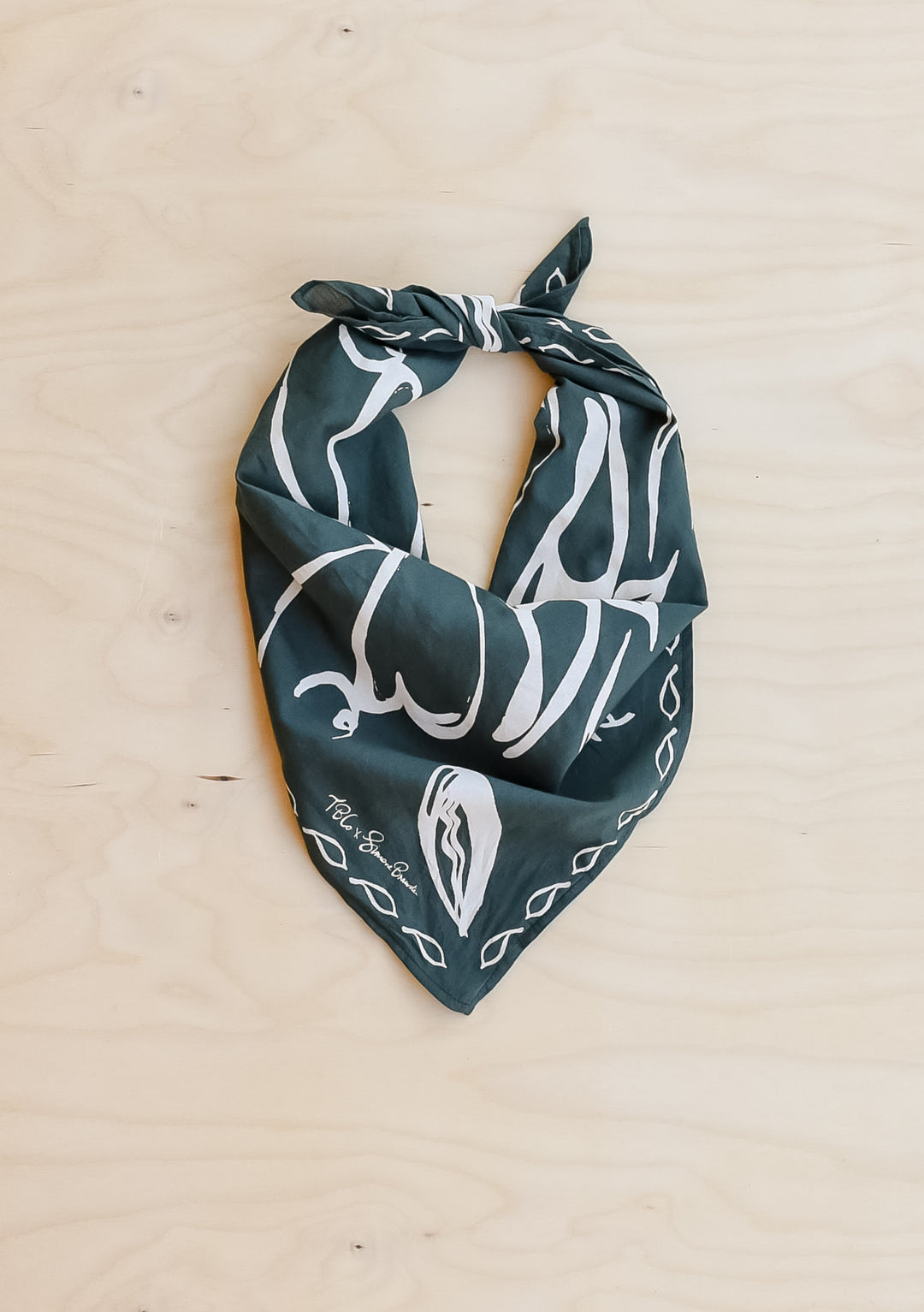 The Artist Series TBCo x Simone Brewster Cotton Scarf, dark green with a white hand-drawn pattern, folded and knotted, displayed on a wood-grain surface.