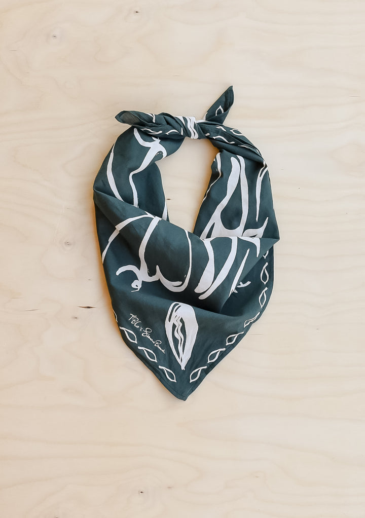 The Artist Series TBCo x Simone Brewster Cotton Scarf, dark green with a white hand-drawn pattern, folded and knotted, displayed on a wood-grain surface.