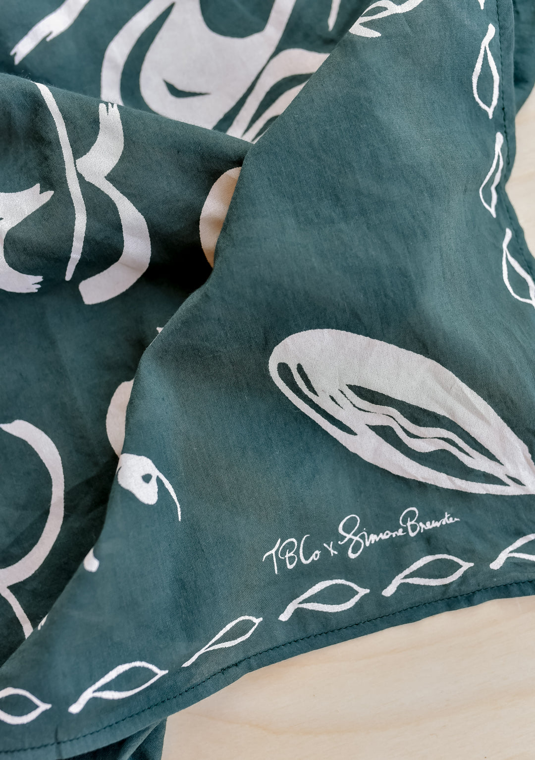 The Artist Series TBCo x Simone Brewster Cotton Scarf features dark green fabric with white hand-drawn fish motifs and a leaf border, signed by the artist.