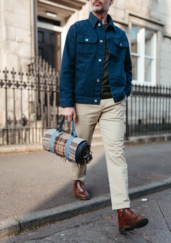 Person holding TBCo Recycled Picnic Carrier in plaid pattern while wearing navy jacket and khaki trousers on city street