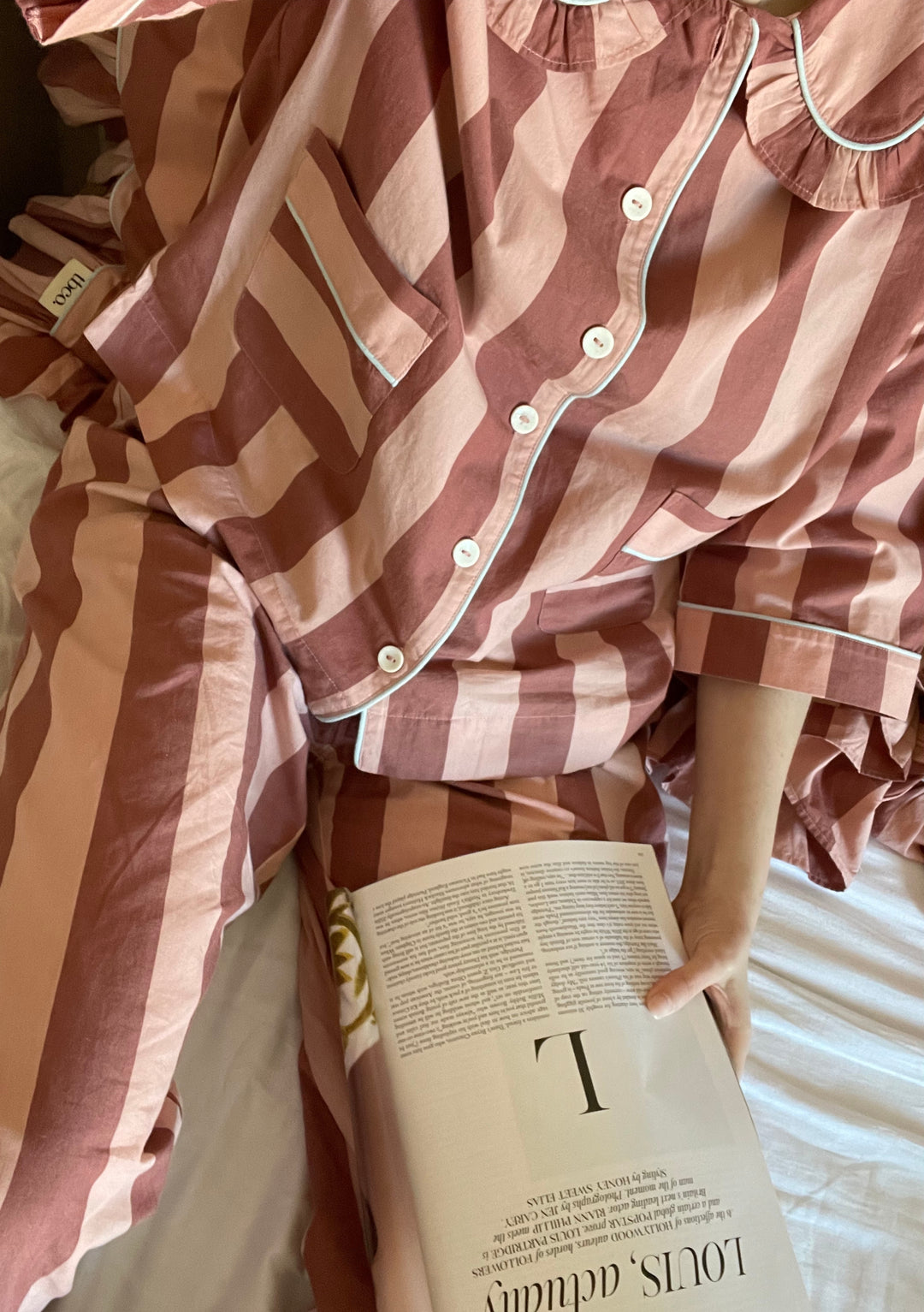 TBCo Cotton Pyjamas in Pink & Red Stripe with ruffle collar and white button details, shown with book while lounging