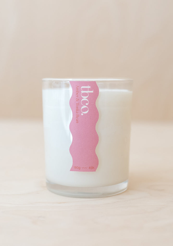 Amber & Tonka Bean Candle in clear glass container with white wax and pink wavy label against beige background