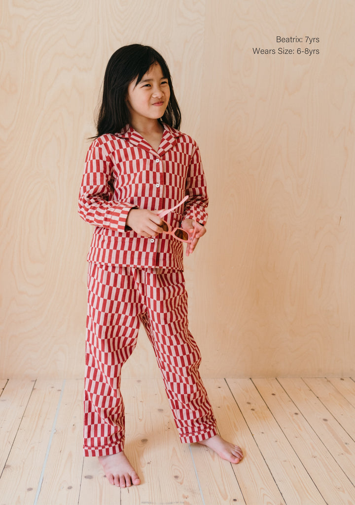 Young model wearing TBCo Cotton Kids Pyjamas in Rose Checkerboard pattern, featuring collared top and matching trousers on wooden floor