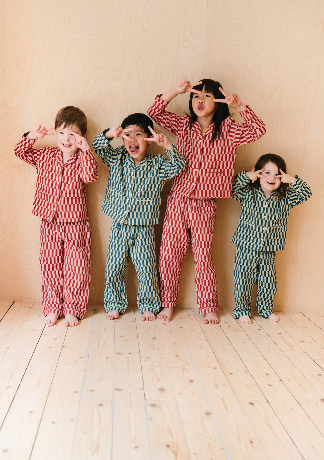 Children wearing TBCo Cotton Kids Pyjamas in Rose Checkerboard and mint green patterns, playfully posing against beige wall