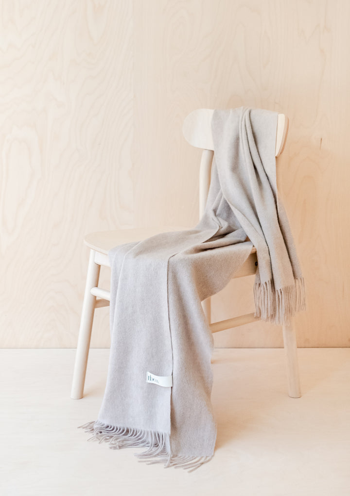 TBCo Cashmere Small Blanket in Oatmeal Melange elegantly draped over white chair, showcasing fringed edges and luxurious texture
