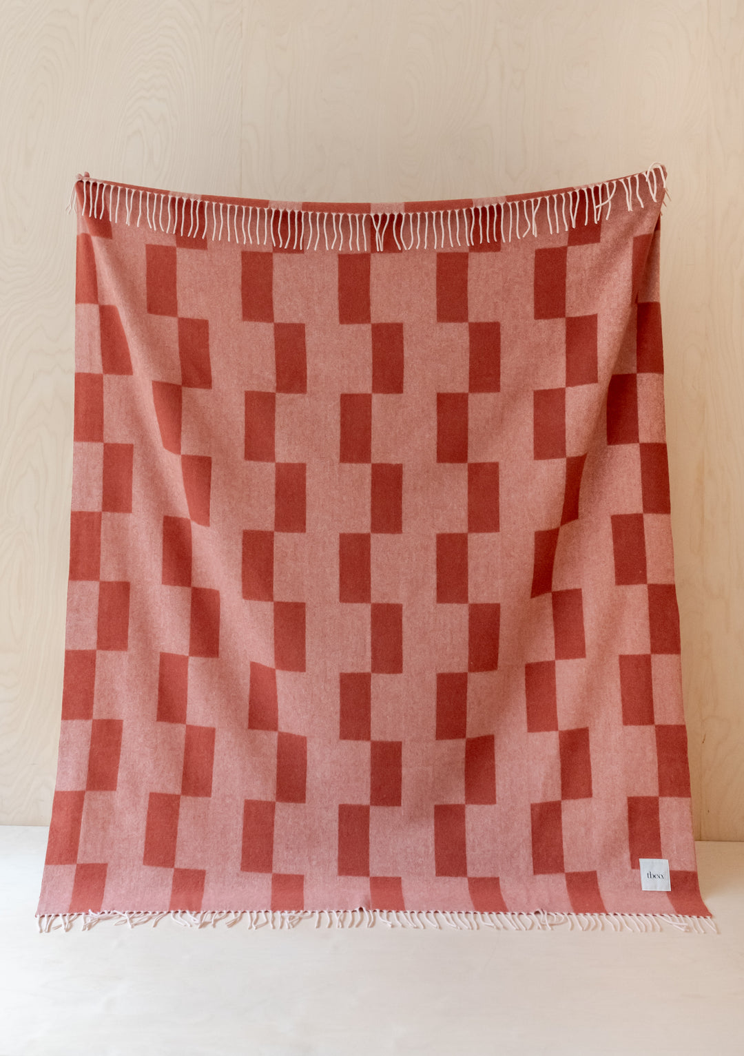 TBCo Lambswool Blanket in Apricot Checker featuring stacked geometric pattern with white fringe against light wood wall