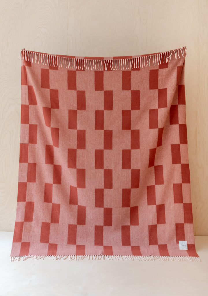 TBCo Lambswool Blanket in Apricot Checker featuring stacked geometric pattern with white fringe against light wood wall