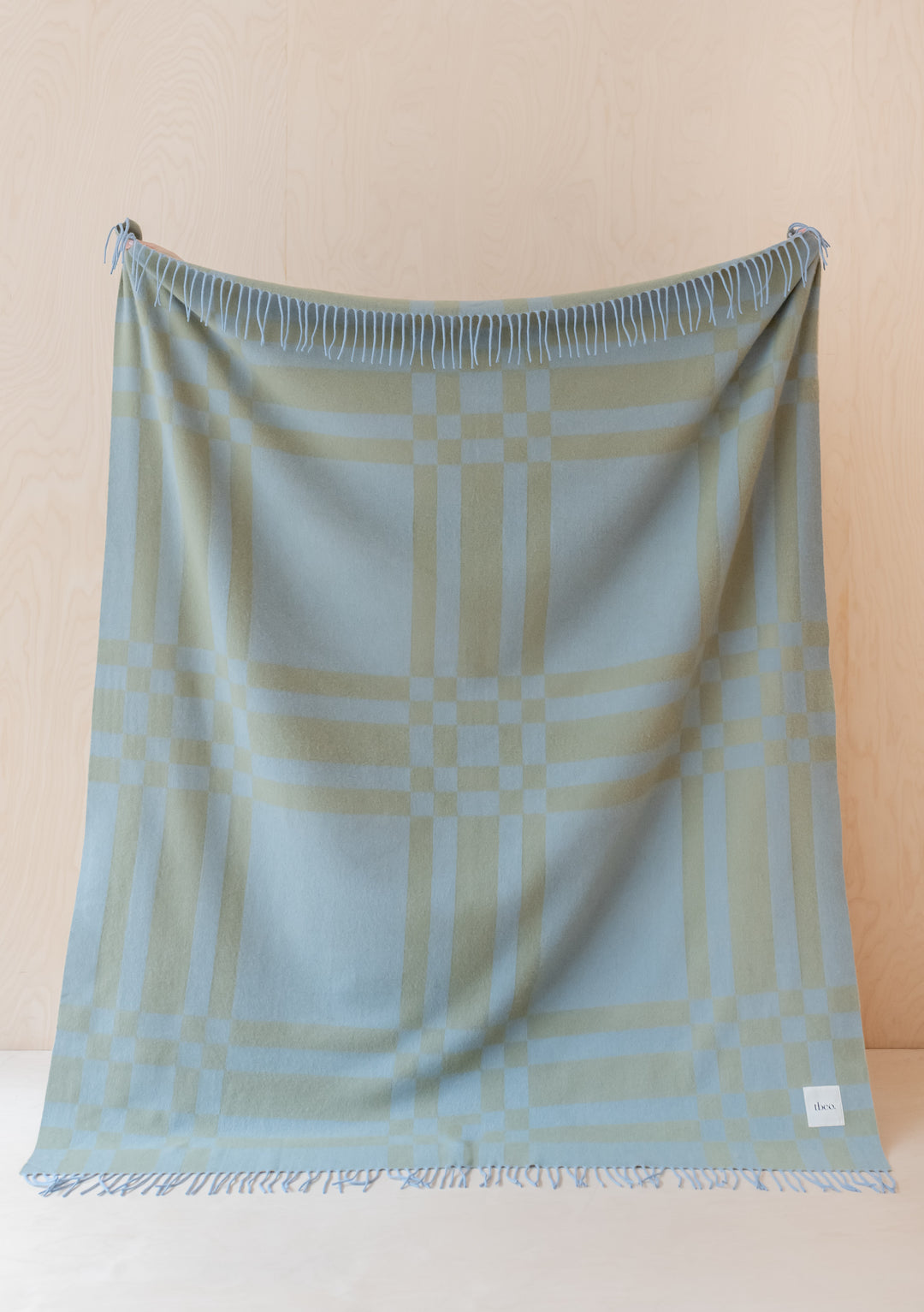 TBCo Lambswool Blanket in blue pillar check pattern displayed vertically against cream wall, showing fringed edges and woven design