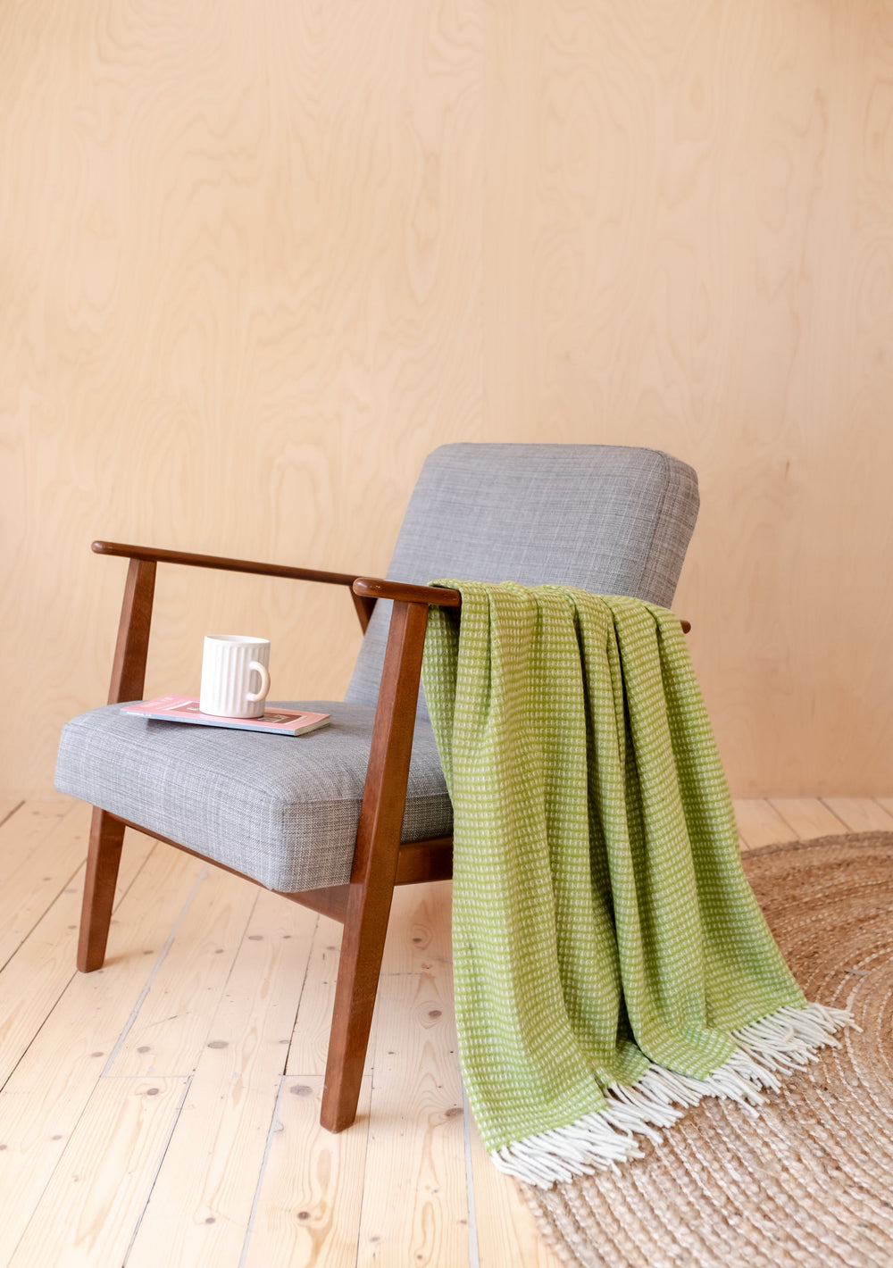 TBCo Recycled Wool Blanket in Green Waffle draped over grey armchair with coffee mug, showcasing textured weave pattern on wooden floor