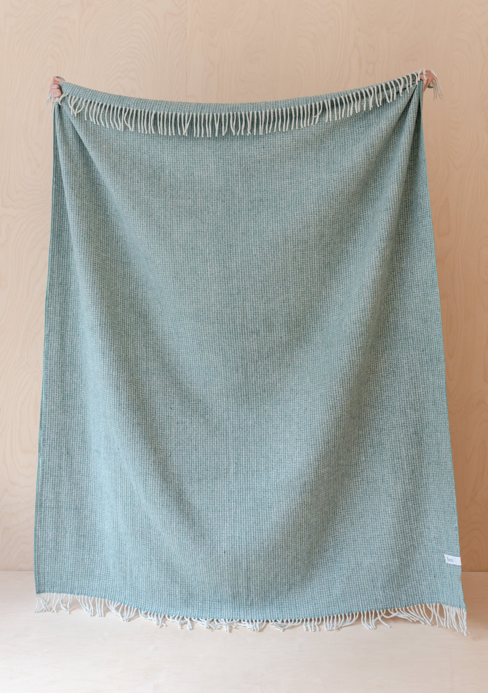 TBCo Recycled Wool Waffle Blanket in sage green with fringed edges, displayed hanging against beige wall showing textured weave