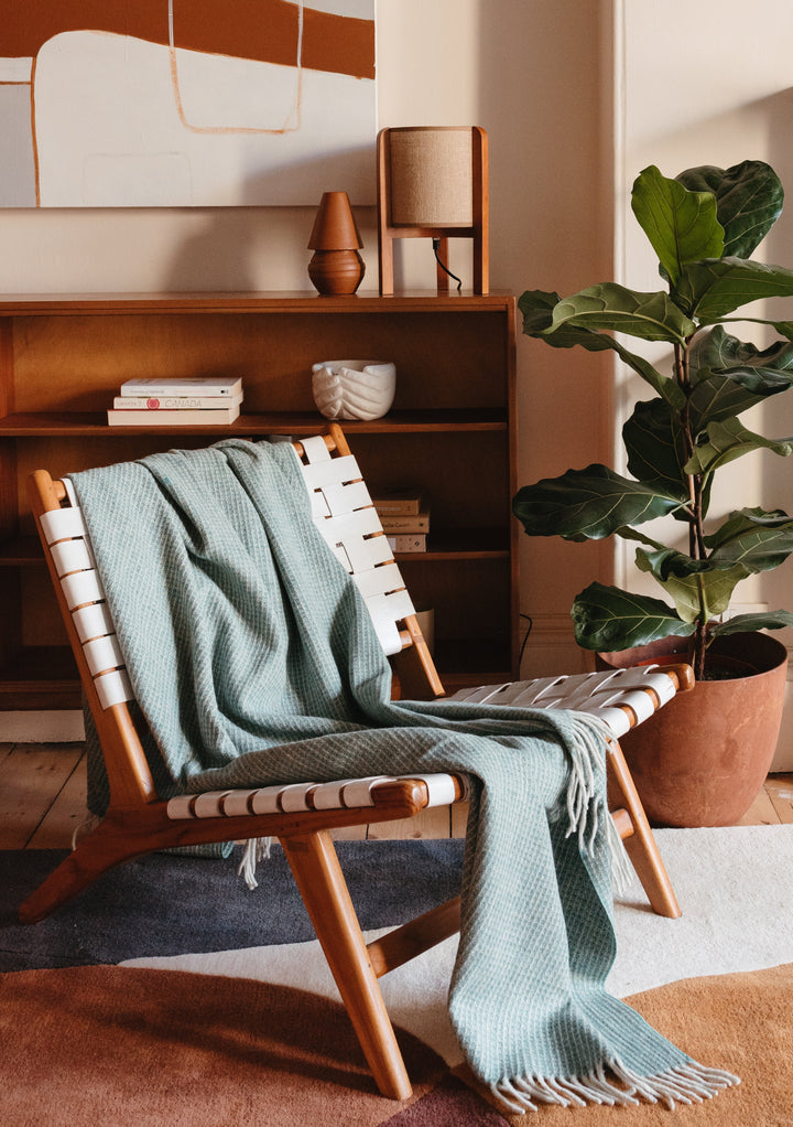 TBCo Recycled Wool Waffle Blanket in Sage draped over wooden lounge chair, styled in modern living room with fiddle leaf plant
