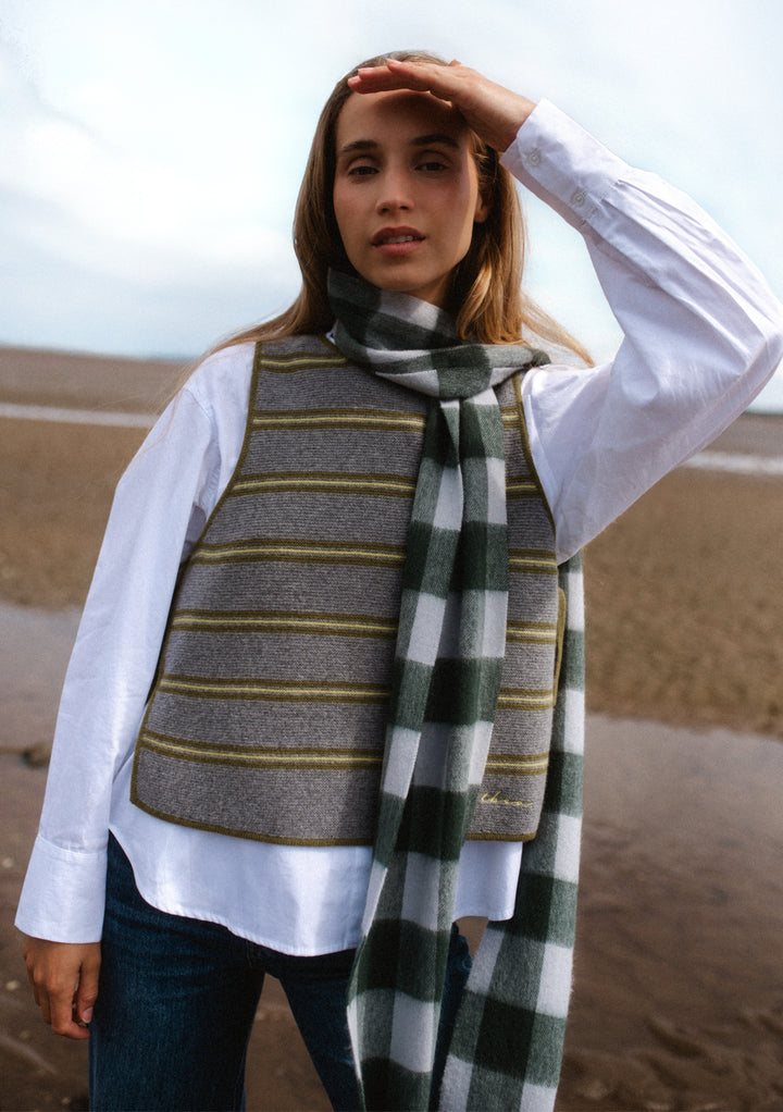 Woman wearing TBCo Lambswool Skinny Scarf in Forest Gingham with white shirt and striped vest on beach setting