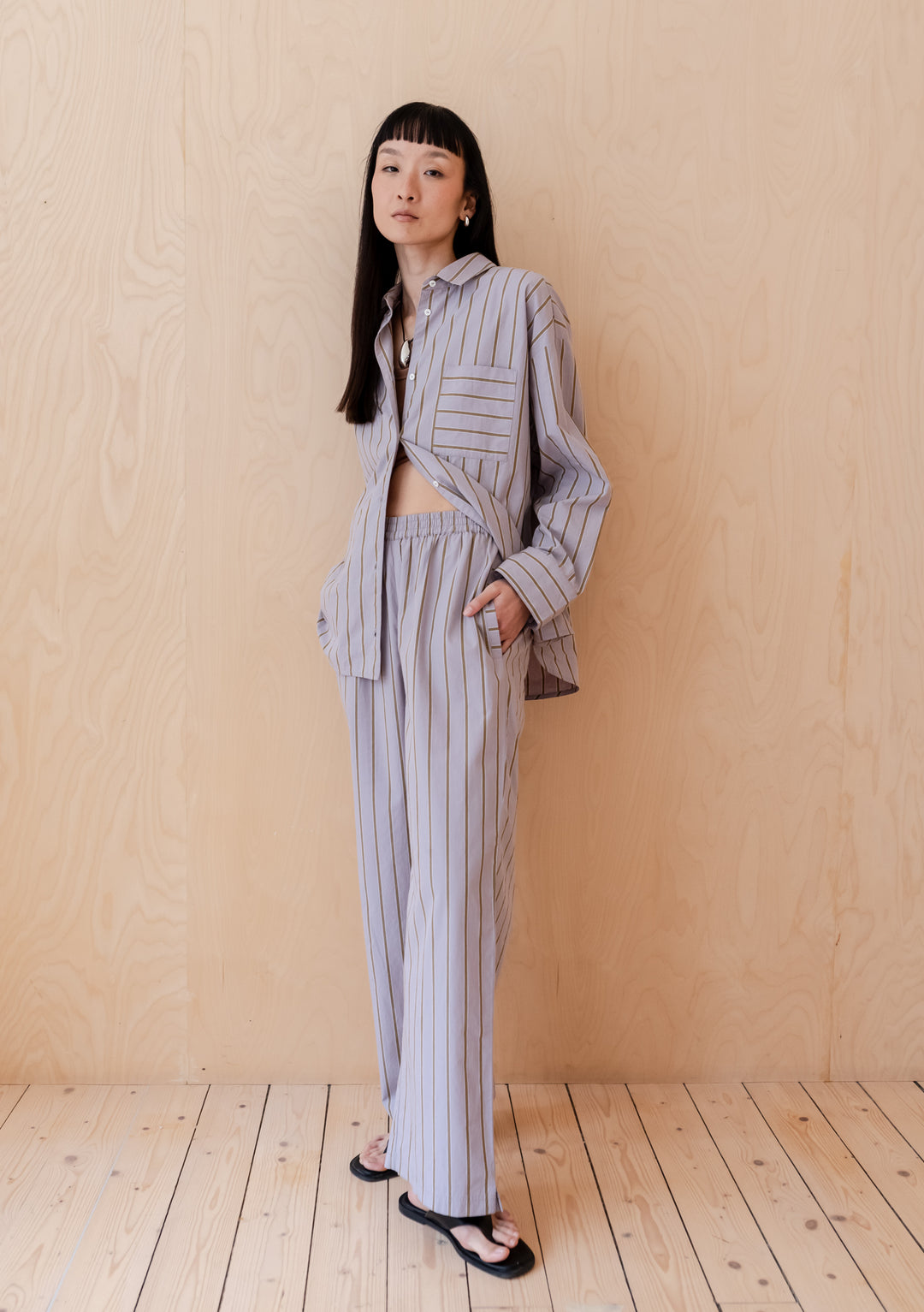 A person wearing TBCo Cotton Trousers in Lilac Stripe with coordinating button-down shirt against wooden background