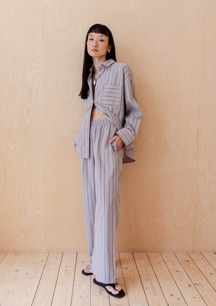 A person wearing TBCo Cotton Trousers in Lilac Stripe with coordinating button-down shirt against wooden background