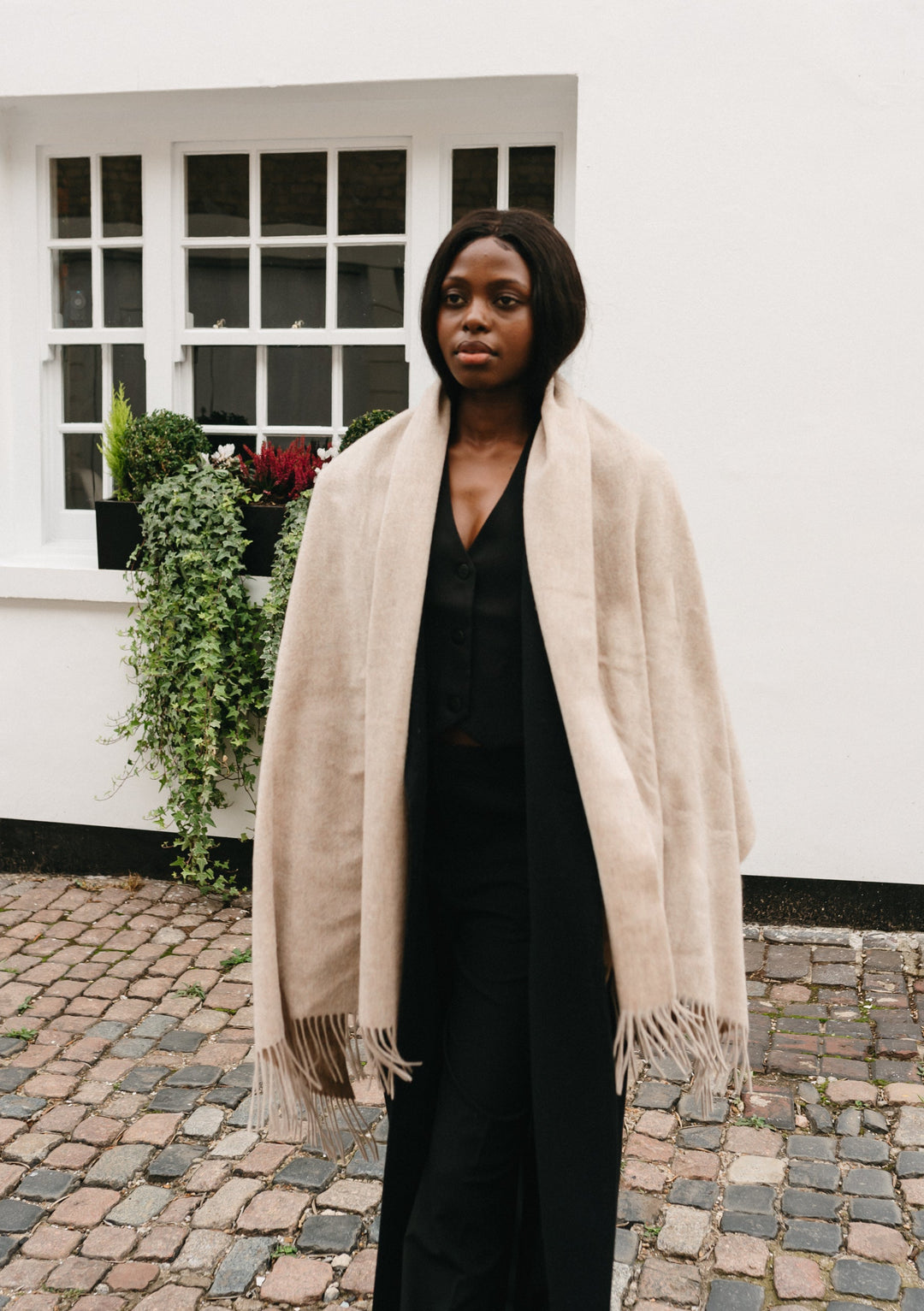 Luxurious TBCo lambswool blanket scarf in oatmeal melange styled with black outfit on cobblestone courtyard with white building
