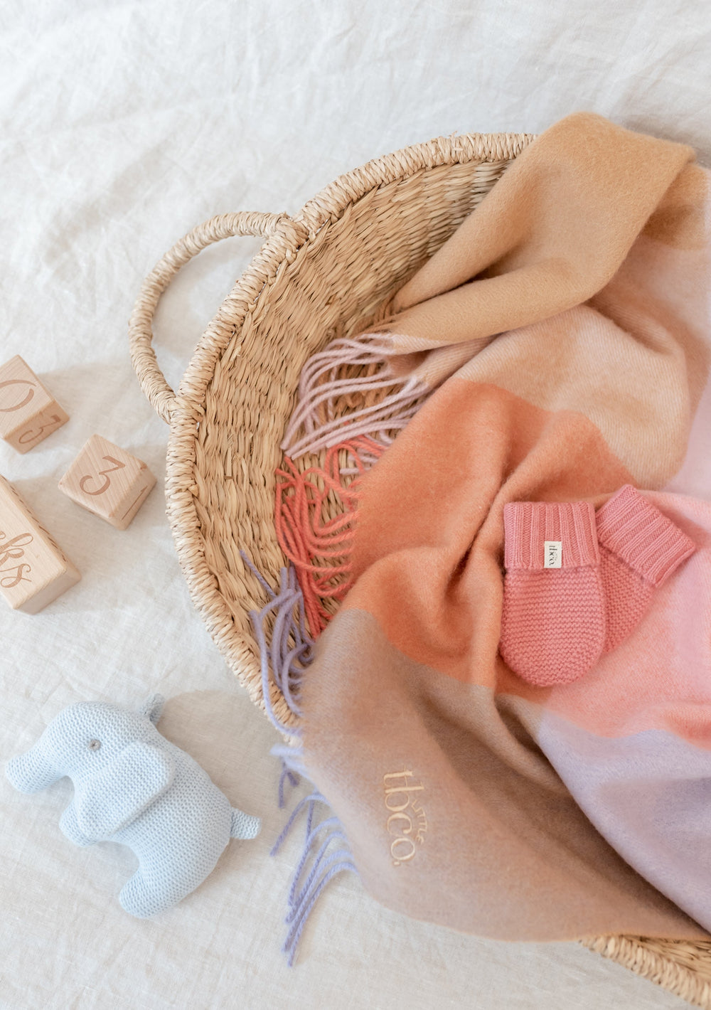 TBCo rose merino wool baby mittens displayed in woven basket with blanket, wooden blocks and knitted elephant toy