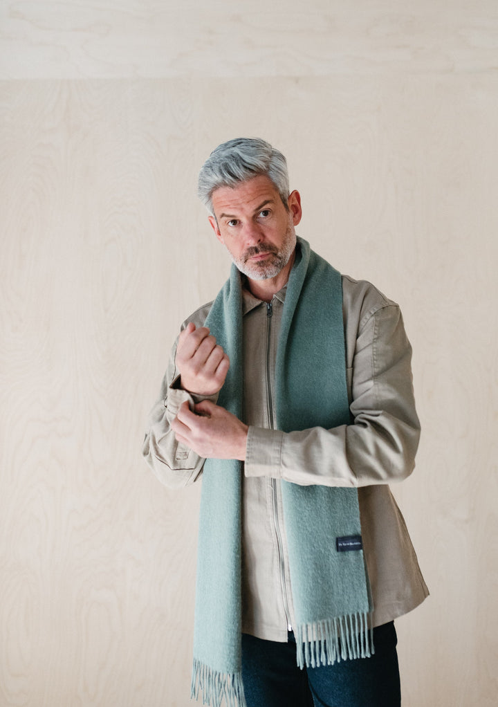 Lifestyle shot of TBCo Mens Lambswool Scarf in Sage Melange styled with beige jacket against neutral background