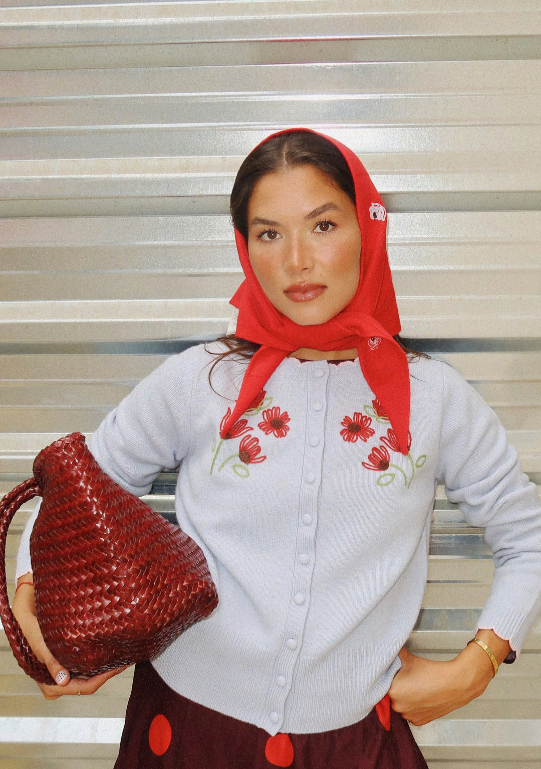 A woman wearing a red headscarf, a Nobody's Child x TBCo Blue Floral Wool Cardigan, and a burgundy skirt holds a woven red bag. She stands before a metallic corrugated backdrop, gazing confidently at the camera.