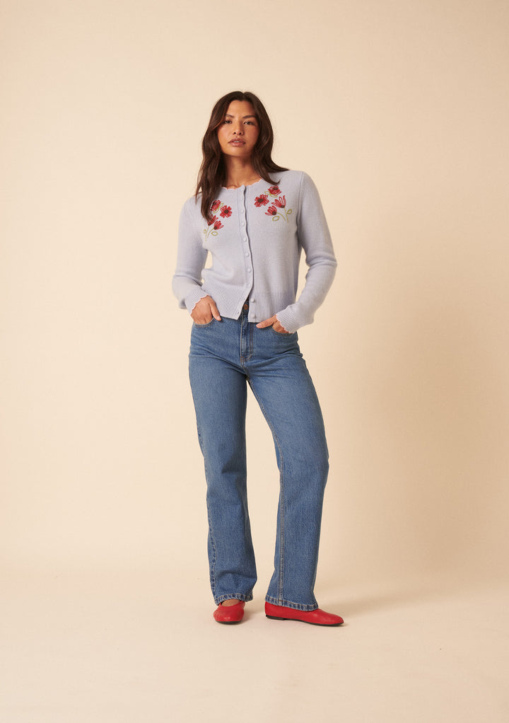 A woman with long brown hair stands against a light beige background, hands in pockets, wearing the Nobody's Child x TBCo Blue Floral Wool Cardigan with blue jeans and red shoes, effortlessly highlighting sustainable knitwear style.