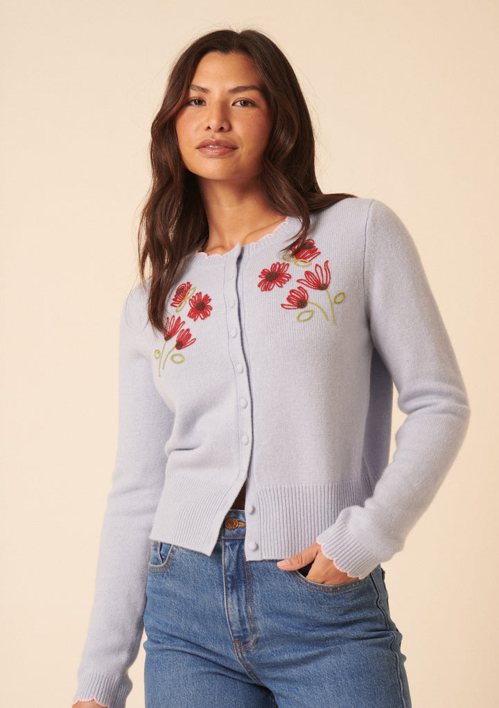 A woman in the Nobody's Child x TBCo Blue Floral Wool Cardigan, featuring light purple tones and red chest embroidery, pairs it with blue jeans against a beige background—showcasing sustainable knitwear elegance.