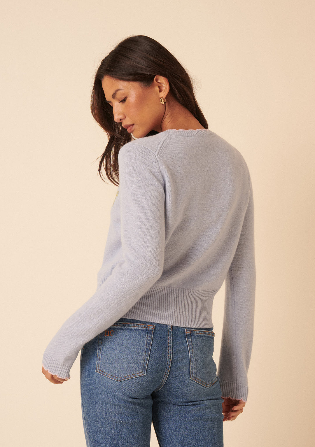 A woman with long brown hair models the Nobody's Child x TBCo Blue Floral Wool Cardigan, paired with blue jeans, standing slightly turned and looking over her shoulder with a beige background behind her.