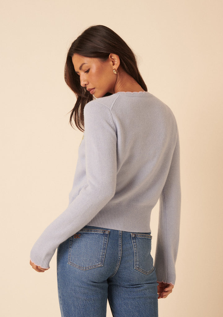 A woman with long brown hair models the Nobody's Child x TBCo Blue Floral Wool Cardigan, paired with blue jeans, standing slightly turned and looking over her shoulder with a beige background behind her.