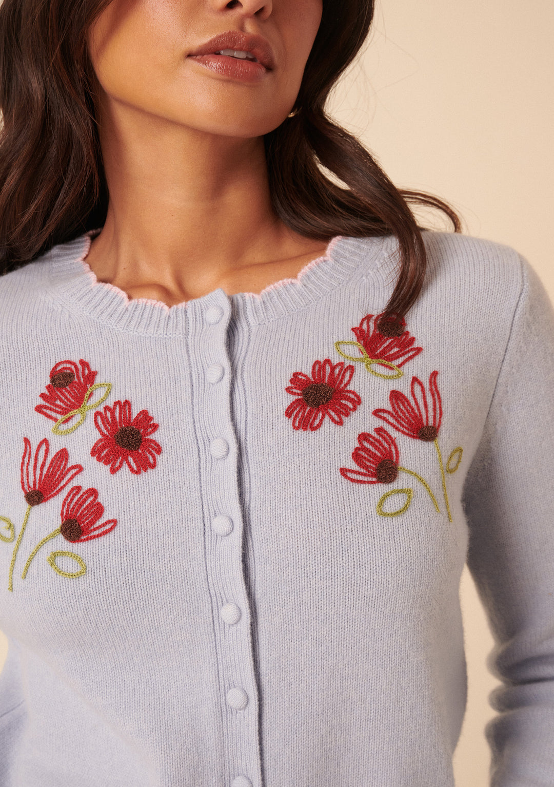 A woman wears the Nobody's Child x TBCo Blue Floral Wool Cardigan, featuring floral embroidery, round buttons, and a scalloped neckline—a charming sustainable knitwear piece.