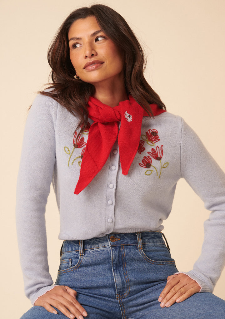 A woman with long dark hair wears the Nobody's Child x TBCo Blue Floral Wool Cardigan, paired with a red scarf and high-waisted blue jeans. She smiles softly, gazing upward, showcasing this sustainable knitwear look.