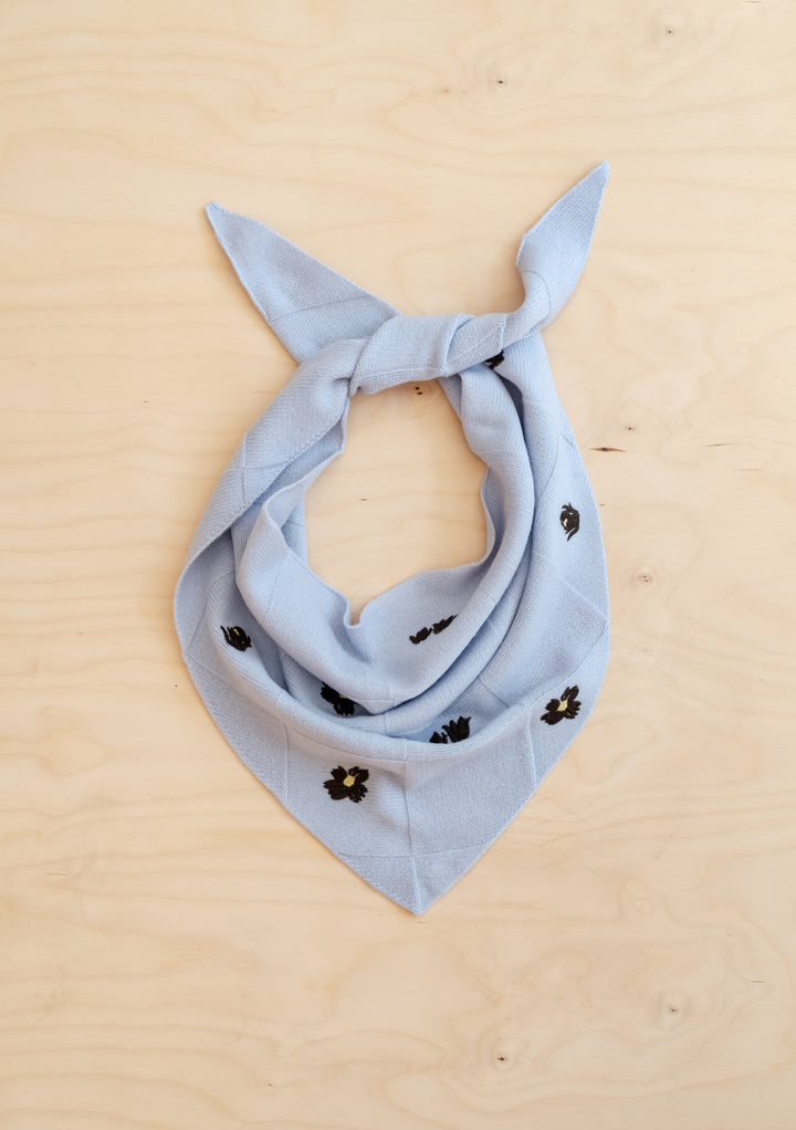 NC x TBCo Blue Floral Wool Triangle Scarf in pale blue merino wool with brown and yellow embroidery detail on wooden surface