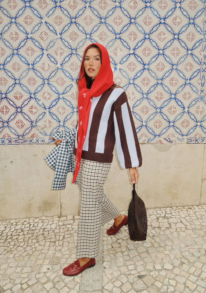 A woman in a red hooded scarf, Nobody's Child x TBCo Blue Stripe Relaxed Wool Jumper, checkered pants, and maroon loafers walks beside a blue and white tiled wall, holding a woven bag and a checkered cloth.