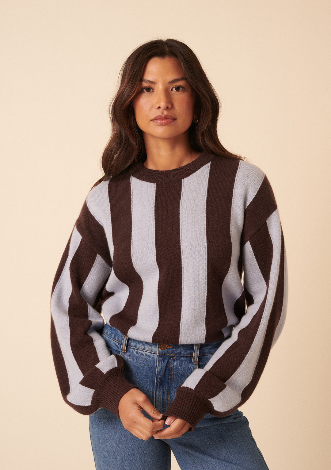 A woman with long, wavy dark hair models the NC x TBCo Blue Stripe Relaxed Wool Jumper and blue jeans, standing against a beige background—an ideal choice for any knitwear capsule.