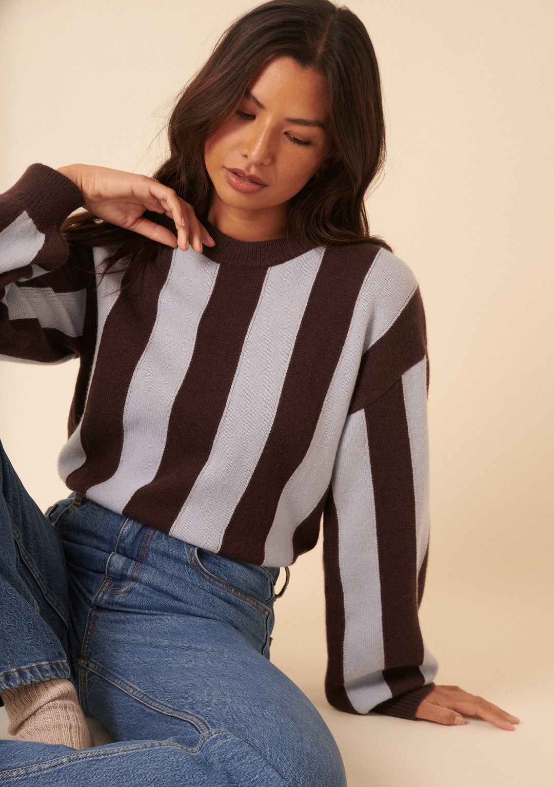 A woman with long dark hair models the NC x TBCo Blue Stripe Relaxed Wool Jumper with blue jeans, sitting with one knee up against a neutral background—a calm, stylish choice for any knitwear capsule wardrobe.