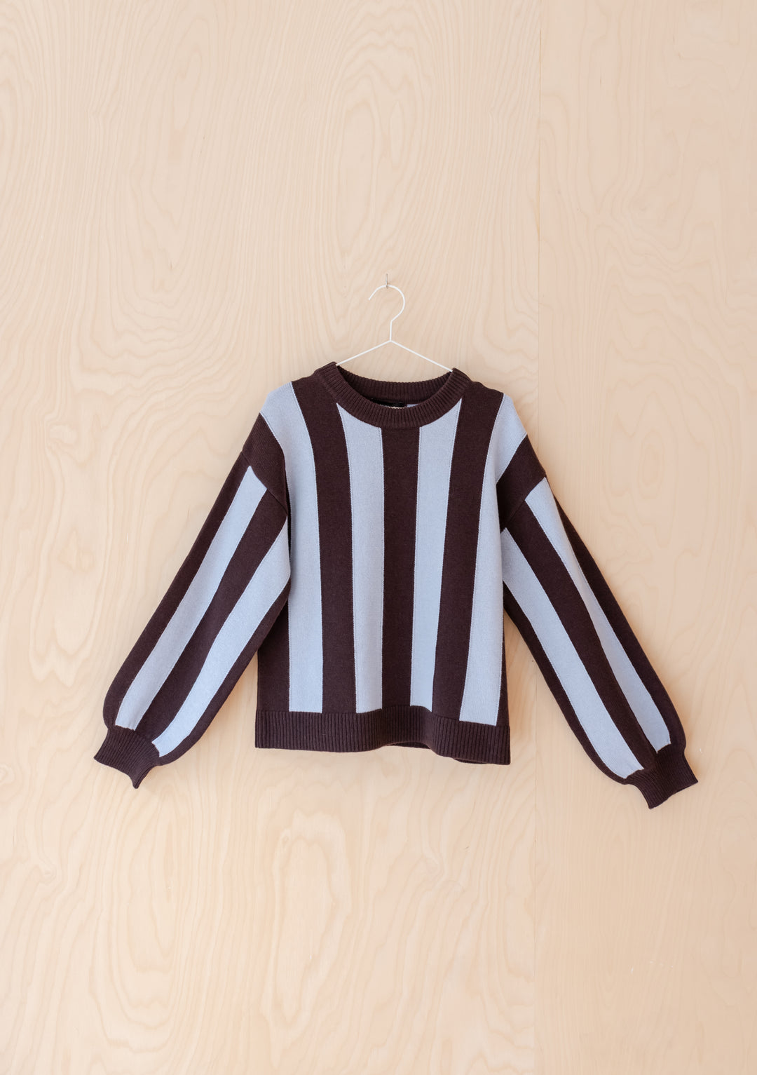 The NC x TBCo Blue Stripe Relaxed Wool Jumper showcases wide blue and brown vertical stripes, long sleeves, and a relaxed fit—displayed on a slim hanger against light wood, it’s a standout piece for any knitwear capsule.