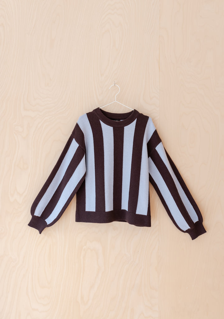 The NC x TBCo Blue Stripe Relaxed Wool Jumper showcases wide blue and brown vertical stripes, long sleeves, and a relaxed fit—displayed on a slim hanger against light wood, it’s a standout piece for any knitwear capsule.