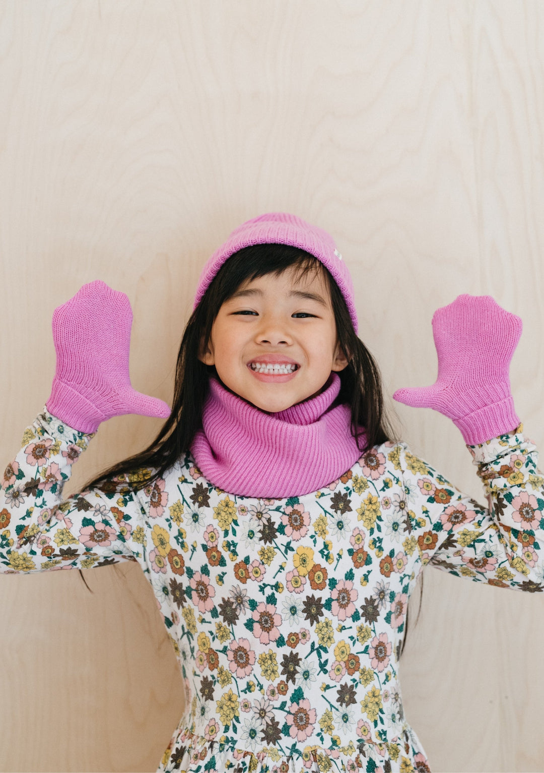 Child wearing TBCo Merino Wool Kids Neck Warmer in Pink with matching beanie and mittens, paired with floral dress