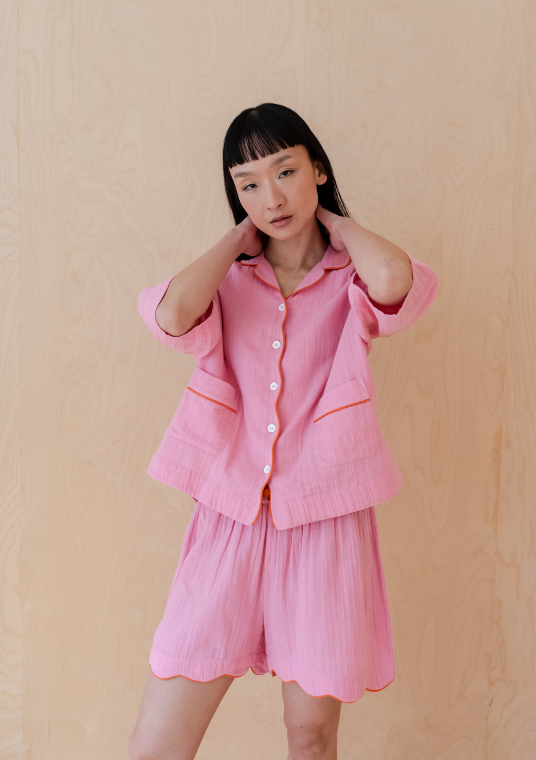Pink cotton pyjama set from TBCo with scalloped edges and orange piping, showcasing elegant loungewear design on beige backdrop