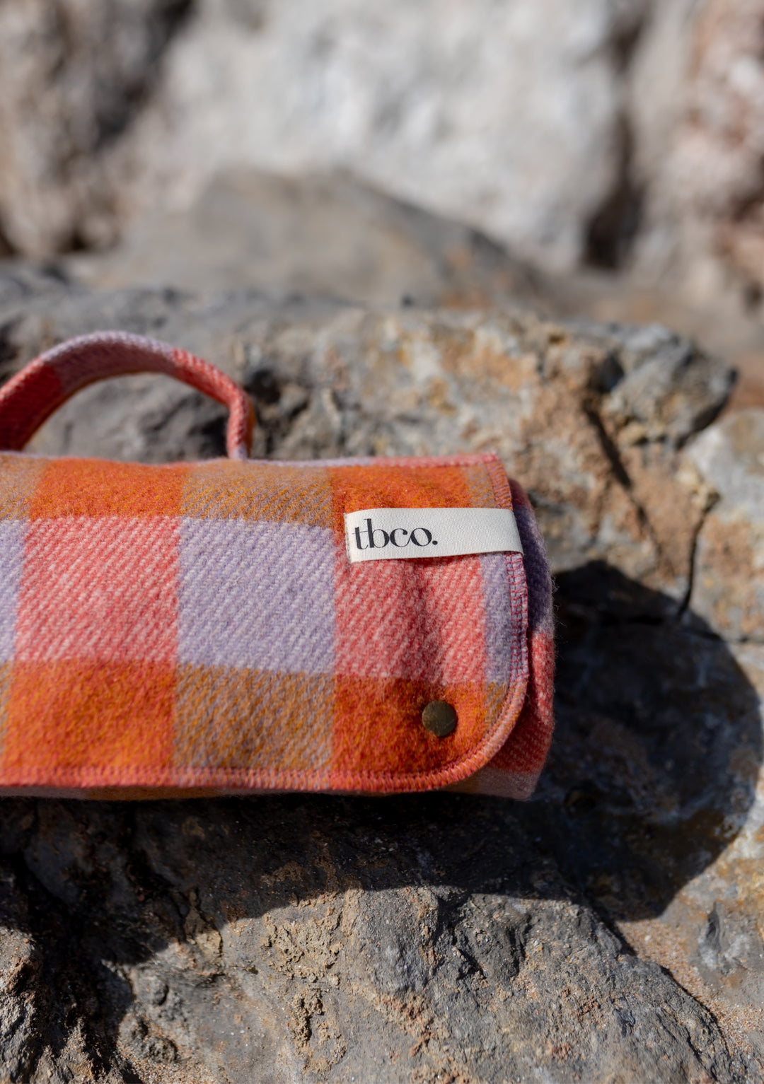 Rolled TBCo recycled wool picnic blanket in orange gingham pattern with lilac accents, featuring carrying handle and popper closure