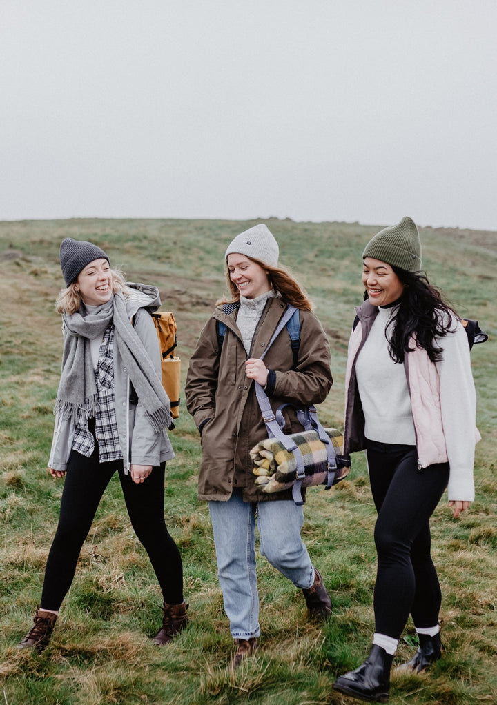 Three friends hiking outdoors carrying TBCo Recycled Picnic Carrier with Shoulder Strap, wearing winter clothing on grassy hillside