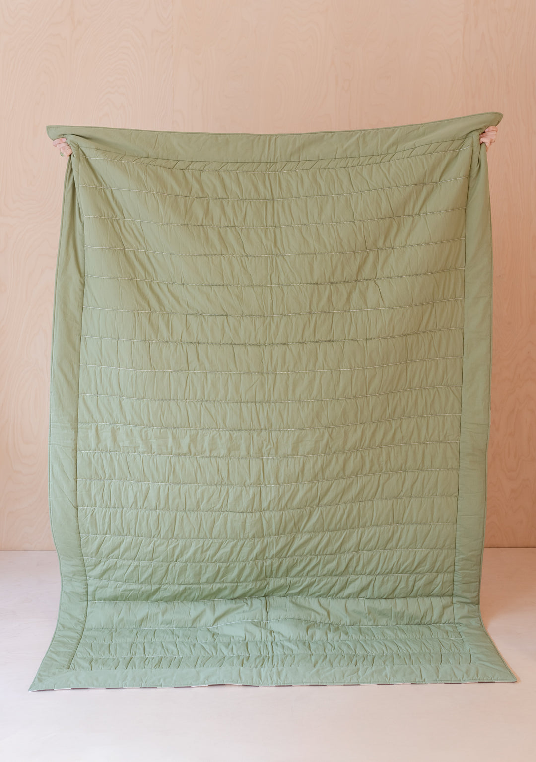 TBCo Celestial Checkerboard quilted cotton throw blanket in sage green, shown from reverse solid side, hung vertically against pink wall